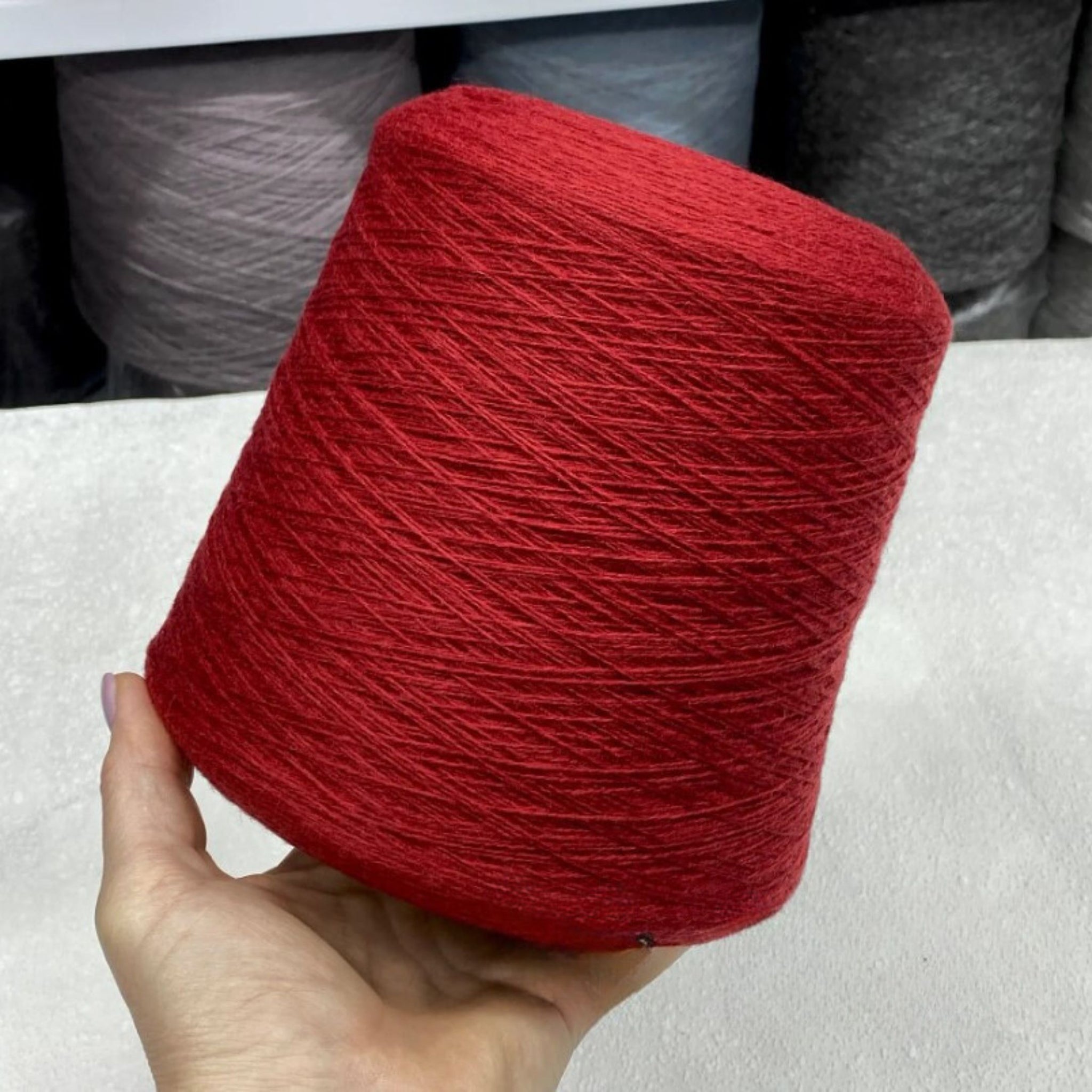 Red yarn ball held by a hand with blurred spools of yarn in the background