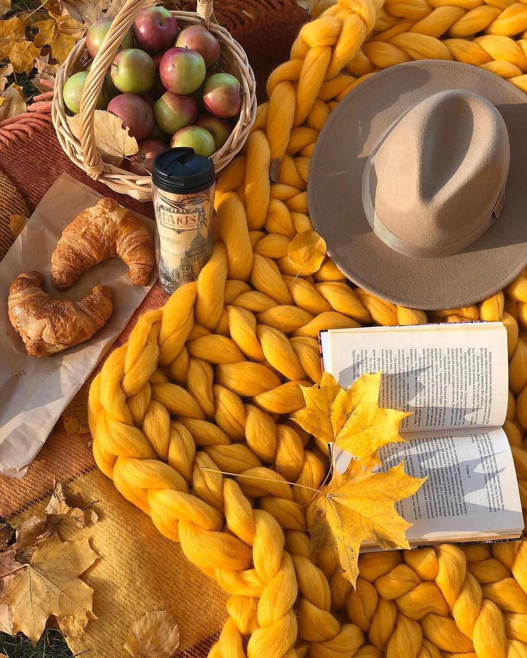 Yellow textured blanket with a basket of apples, croissant, book, and hat on a wooden surface.