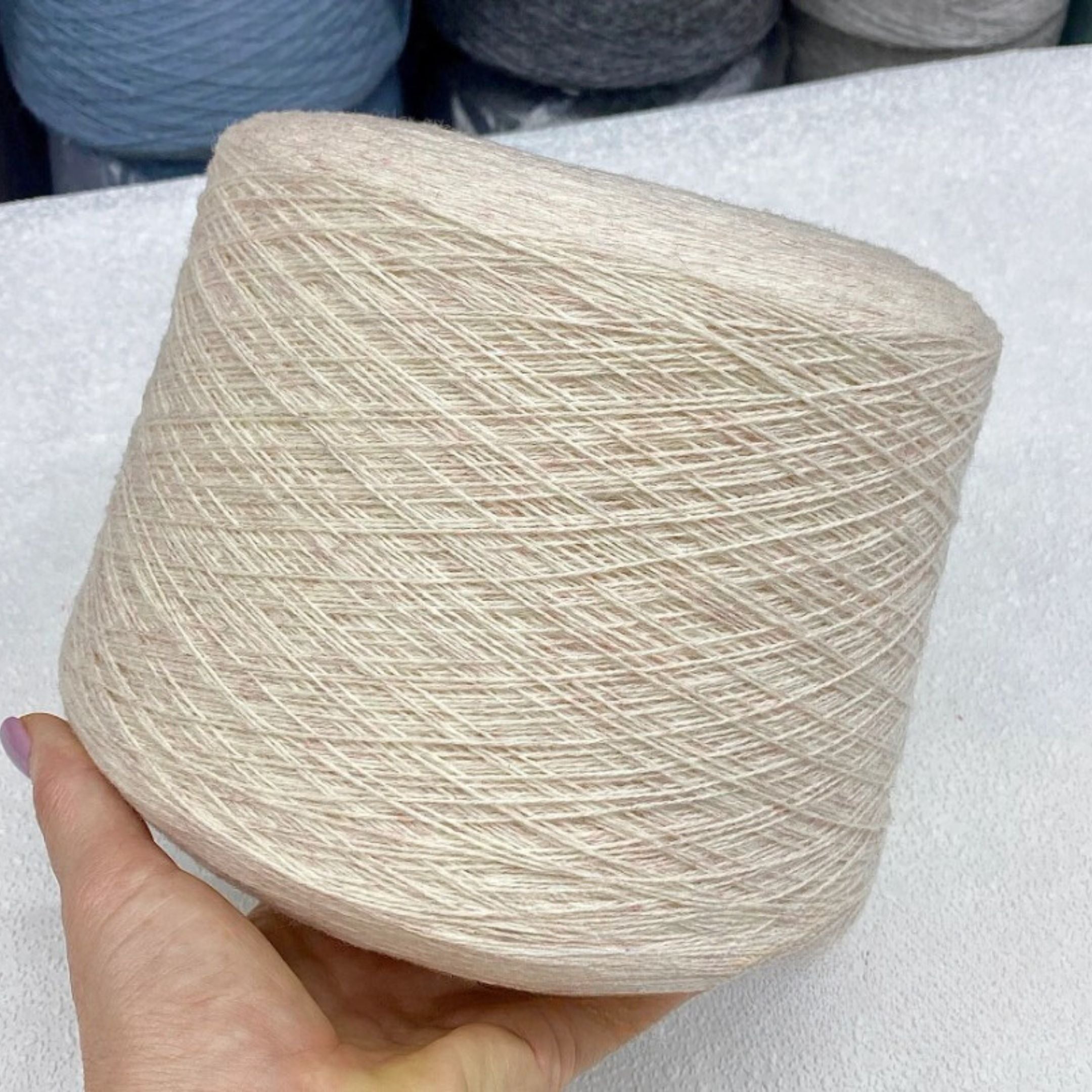 Spool of beige yarn held by a hand on a white surface with more yarn spools in the background.