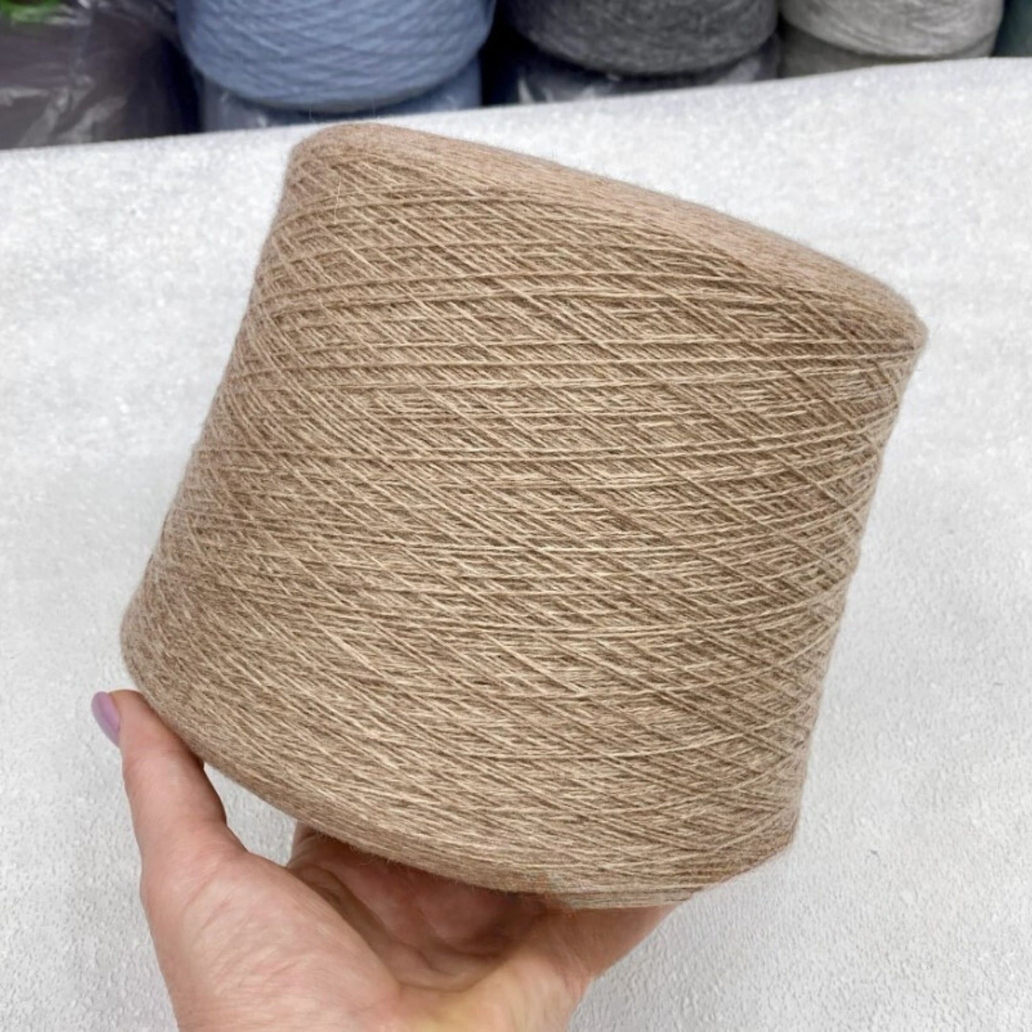 Beige yarn ball held by a hand on a white surface with blurred yarn balls in the background