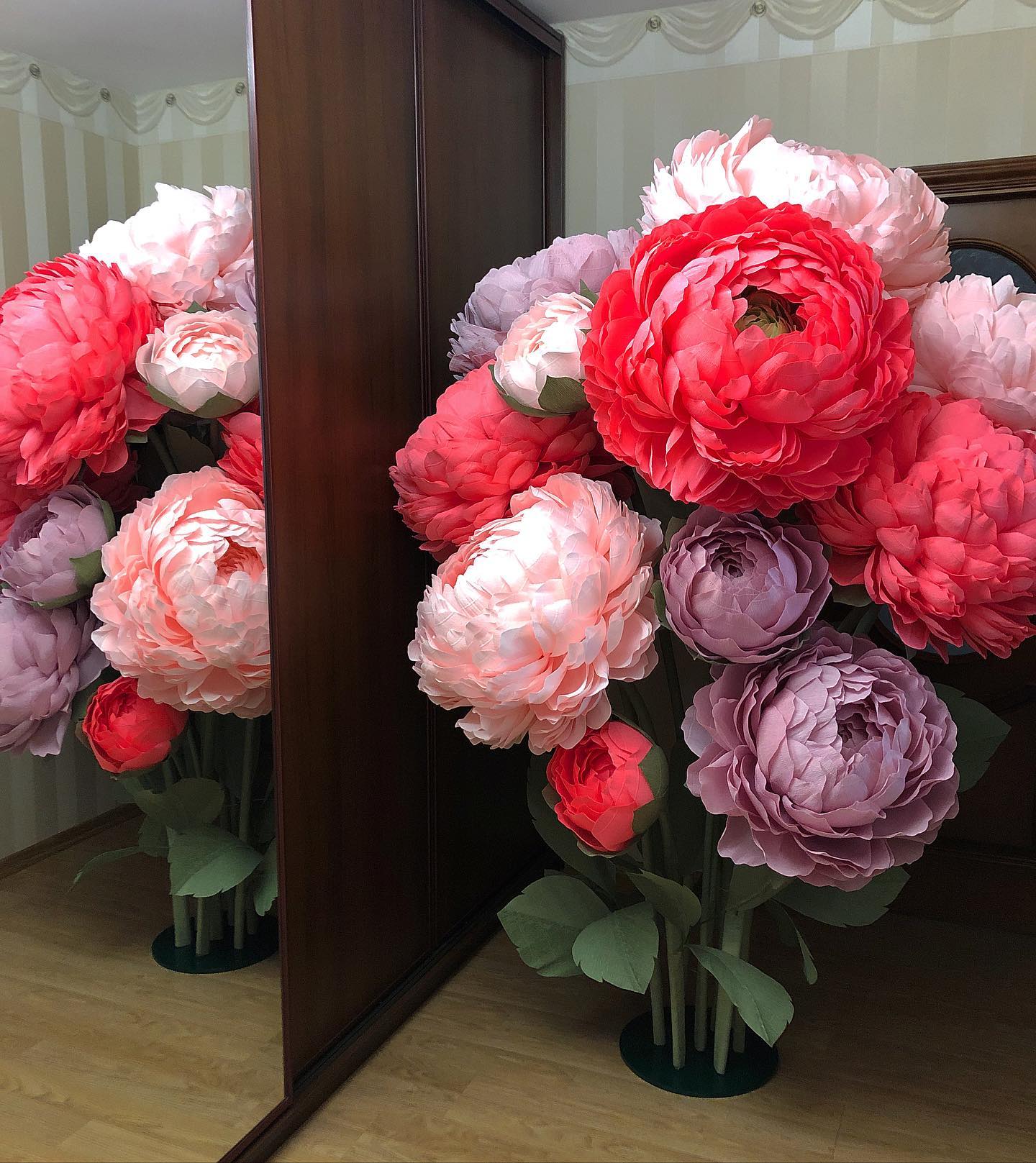 Large giant paper peony flowers in shades of red, pink, lilac and white standing next to a mirror in a modern interior. Handcrafted 65cm peonies creating a stunning wedding photo zone backdrop.