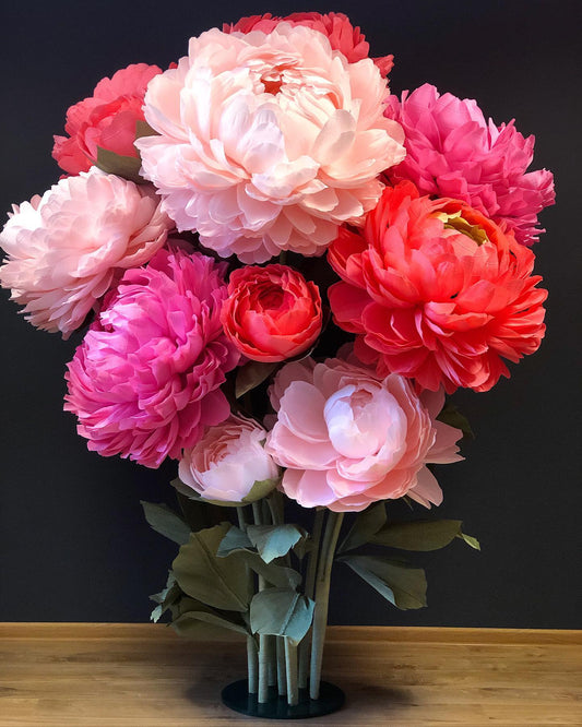 Large bouquet of giant paper peony flowers in soft pink, hot pink, coral and cream shades standing on a wooden table against a dark background. Handmade 65cm crepe paper peonies perfect for wedding decor, photo zones and home decoration.