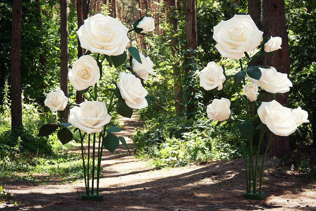 Enchanting giant white foam roses in forest path setting, oversized handmade foam blooms for Alice in Wonderland outdoor wedding backdrop
