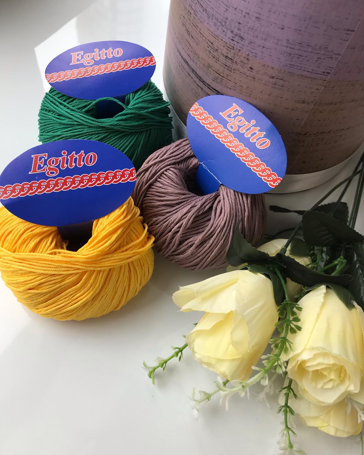 Three balls of yarn in green, yellow, and purple with 'Egitto' labels, next to a bouquet of flowers on a white surface.